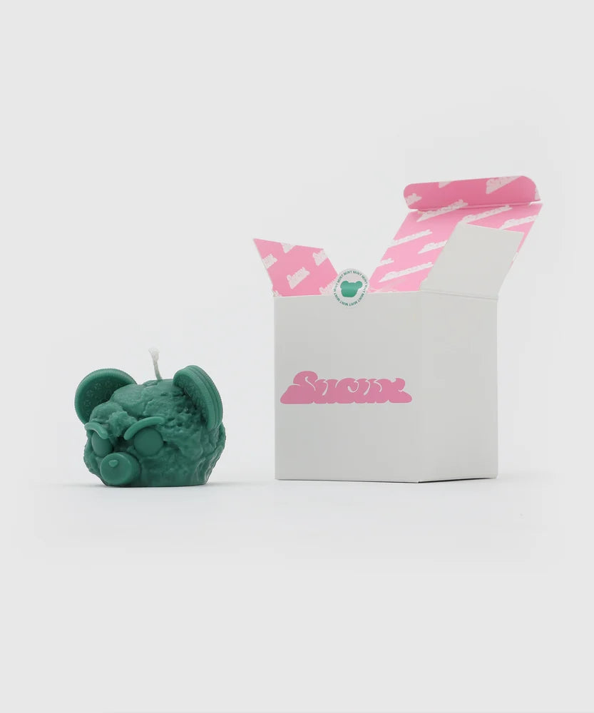 Green bear-shaped candle with cookie ears and pacifier nose next to a white box with pink Sucux logo and custom pink-and-white printed interior – collectible item from the Sucux Cool Collection.
