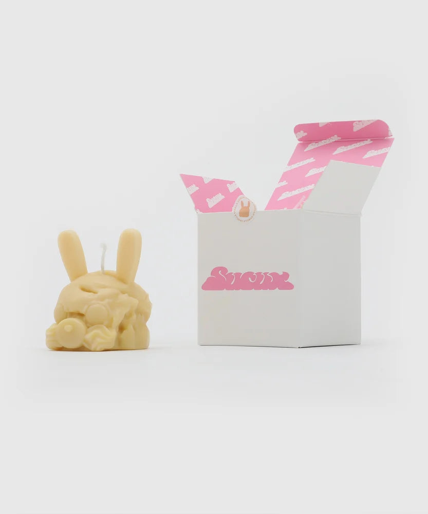“Light yellow bunny-shaped candle with long ears and pacifier nose placed next to a white box with pink Sucux logo and matching pink logo print inside the lid – collectible item from the Sucux Cool Collection.
