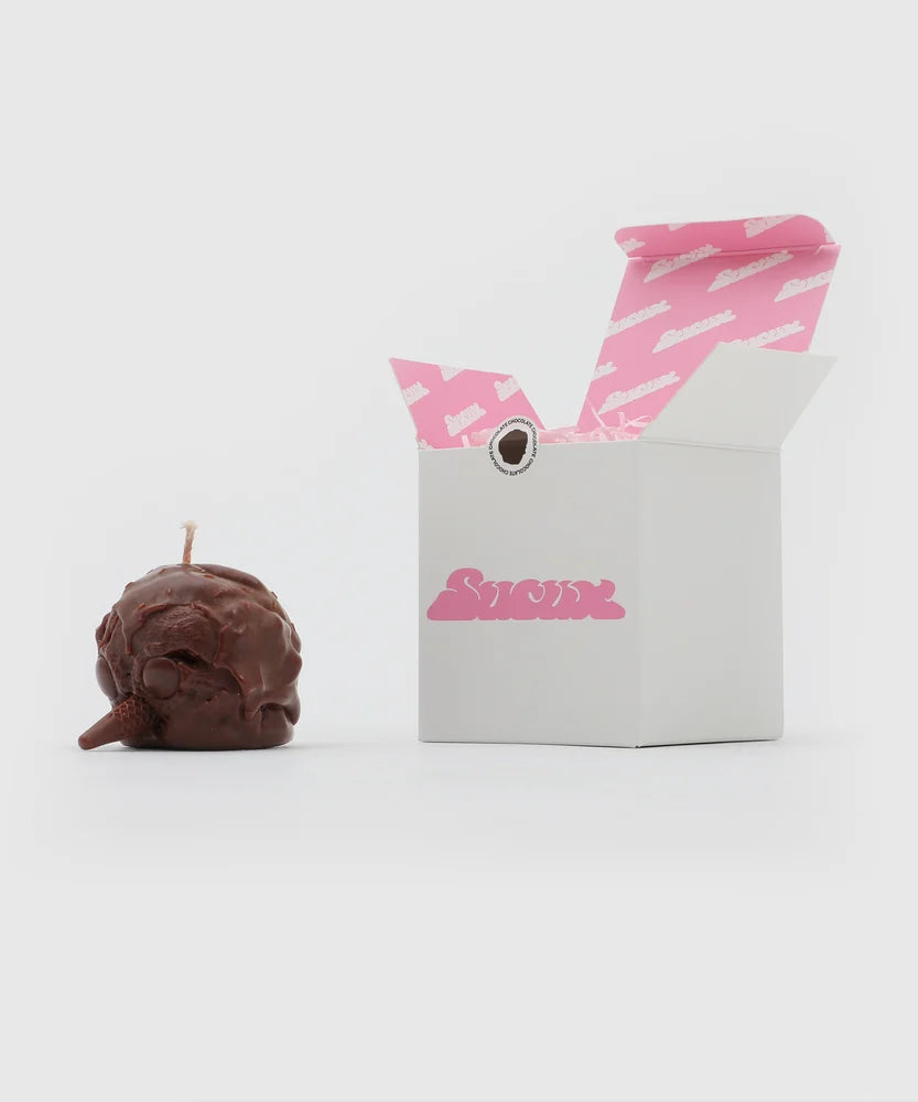 Chocolate-brown penguin-shaped candle with a cone beak and melting scoop texture placed next to a white box with pink Sucux logo and branded pink interior – collectible from the Sucux Cool Collection.