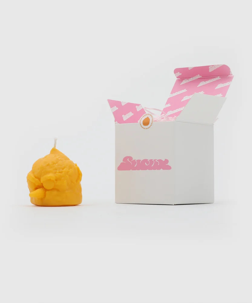 Yellow bird-shaped candle with cone beak and textured scoop look, placed next to a white box featuring the pink Sucux logo and branded pink interior – part of the Sucux Cool Collection.