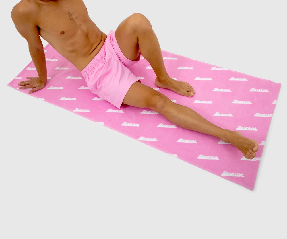 Person lounging on a full-size pink towel with an all-over white Sucux logo print, shown in a relaxed beach or poolside pose – part of the Sucux Cool Collection.