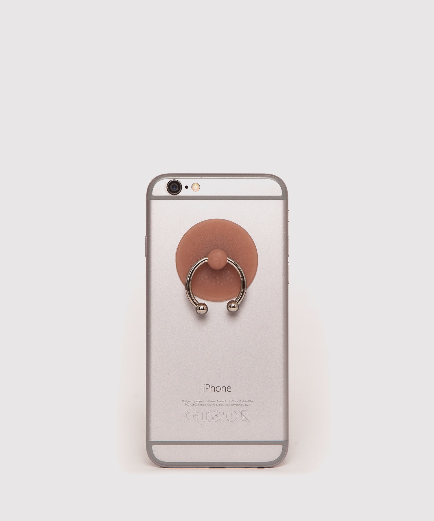 Silver iPhone with a nipple-shaped phone ring holder attached to the back. The holder resembles a realistic nipple with a metal ring, used as a grip or stand for the phone.
