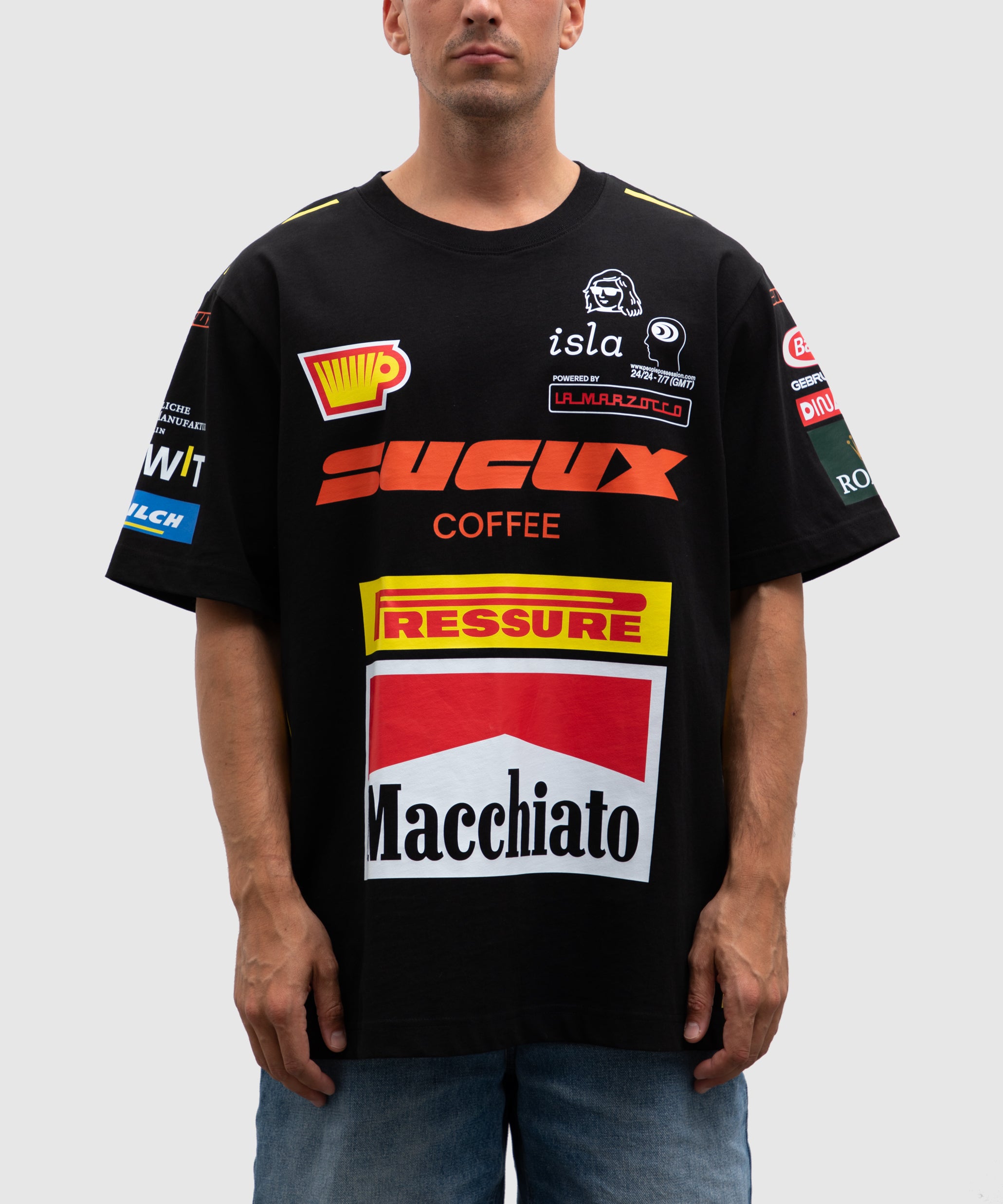 Black oversized SUCUX COFFEE T-shirt inspired by racing and sponsor aesthetics. The front features bold coffee-related graphics and logos including "Macchiato," "Pressure," and "La Marzocco," alongside other playful faux sponsor patches in red, yellow, white, and blue.