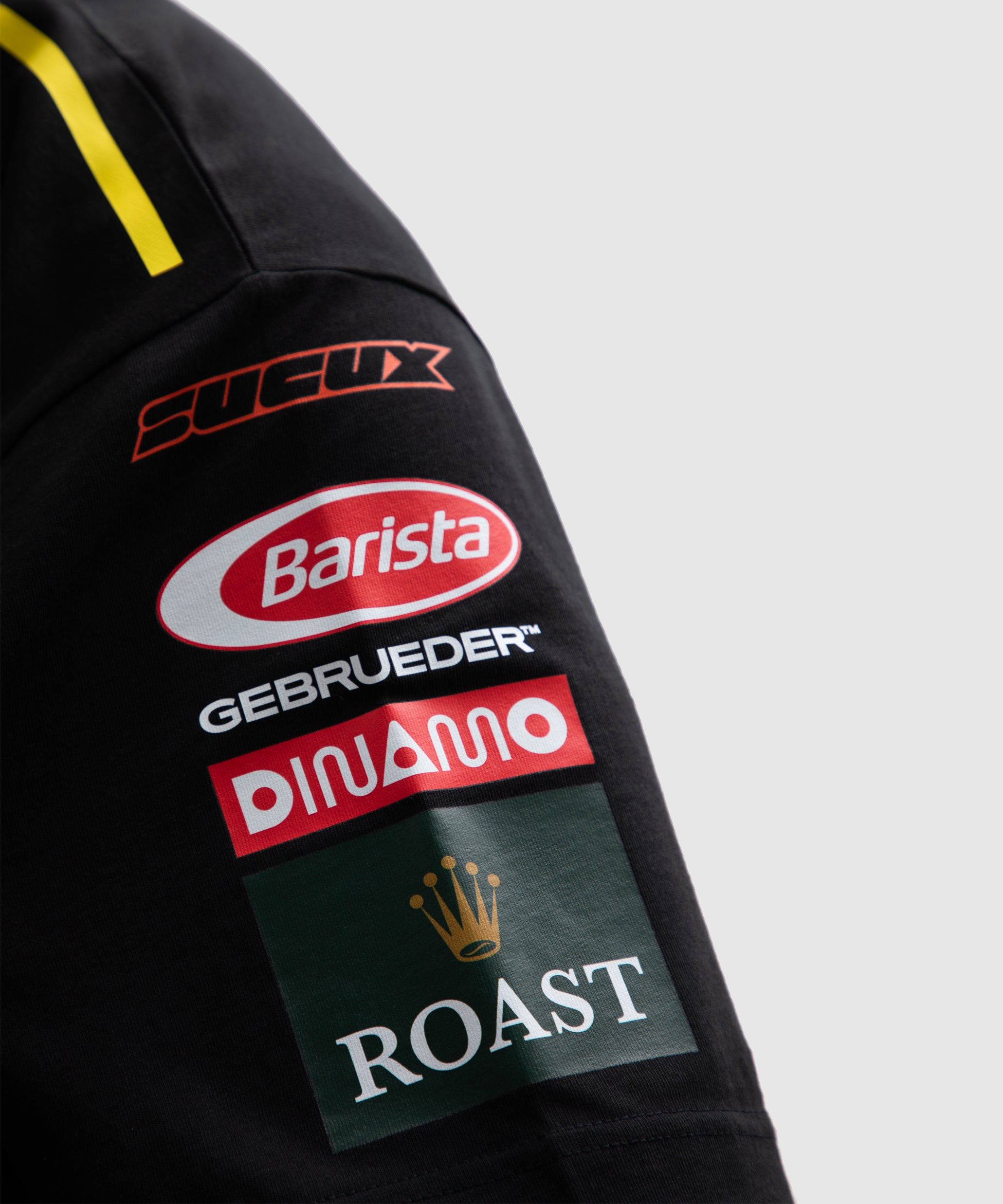 Close-up of the left sleeve of a black SUCUX COFFEE T-shirt, showing mock sponsor-style logos including “SUCUX,” “Barista,” “GEBRUEDER,” “DINAMO,” and “ROAST” with a crown icon—designed in the style of racing team patches.