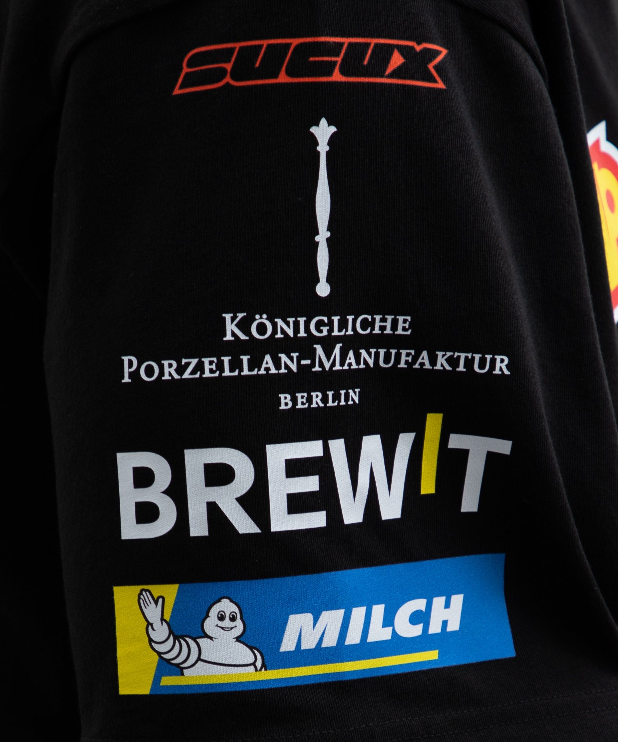 Close-up of the right sleeve of a black SUCUX COFFEE T-shirt, featuring parody sponsor-style logos including “SUCUX,” the Königliche Porzellan-Manufaktur Berlin emblem, “BREWIT,” and a mock Michelin-style “MILCH” patch with a waving cartoon figure.