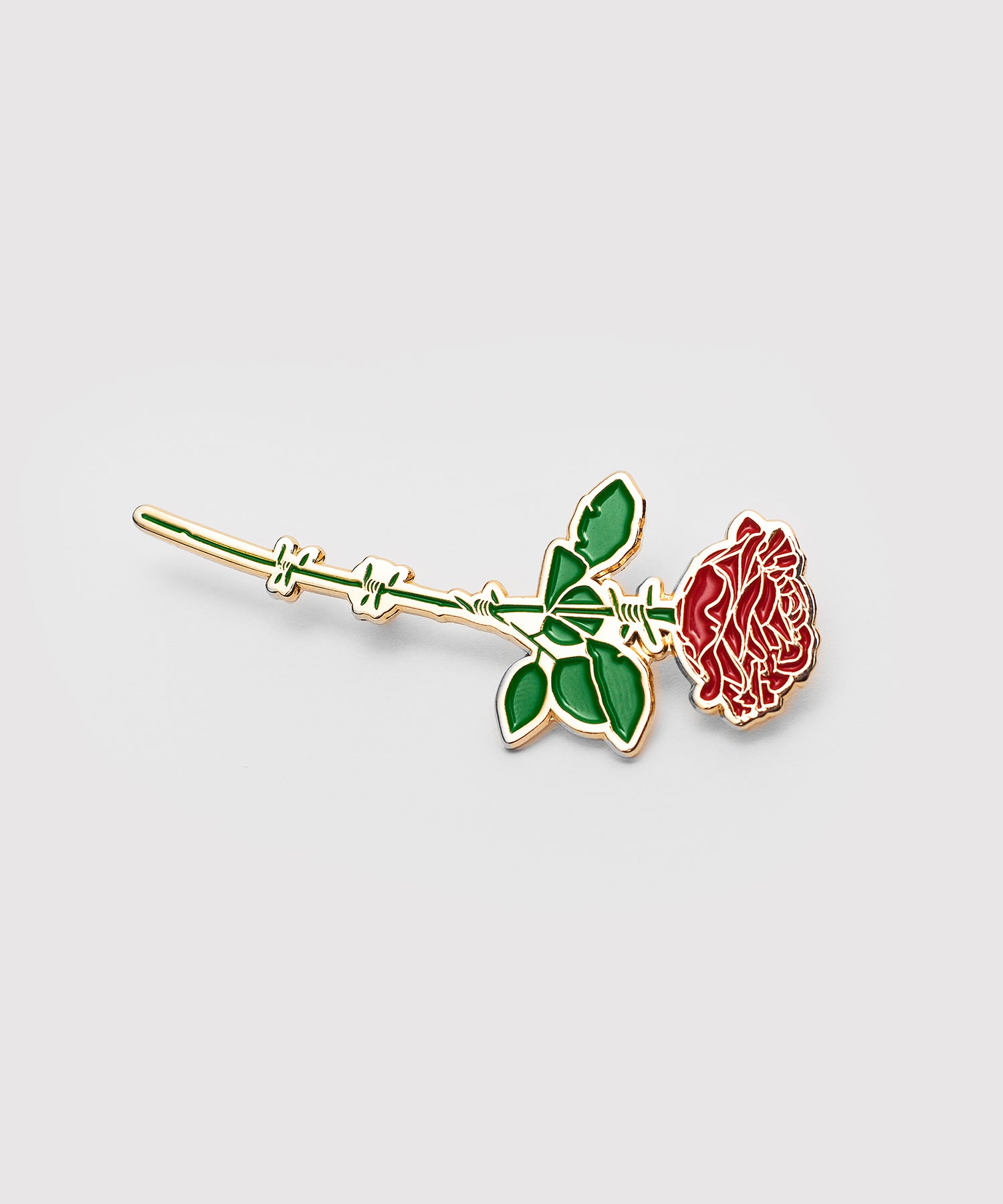 A gold enamel pin shaped like a red rose with green leaves. The stem of the rose is designed as barbed wire, blending delicate floral design with a sharp, contrasting detail.