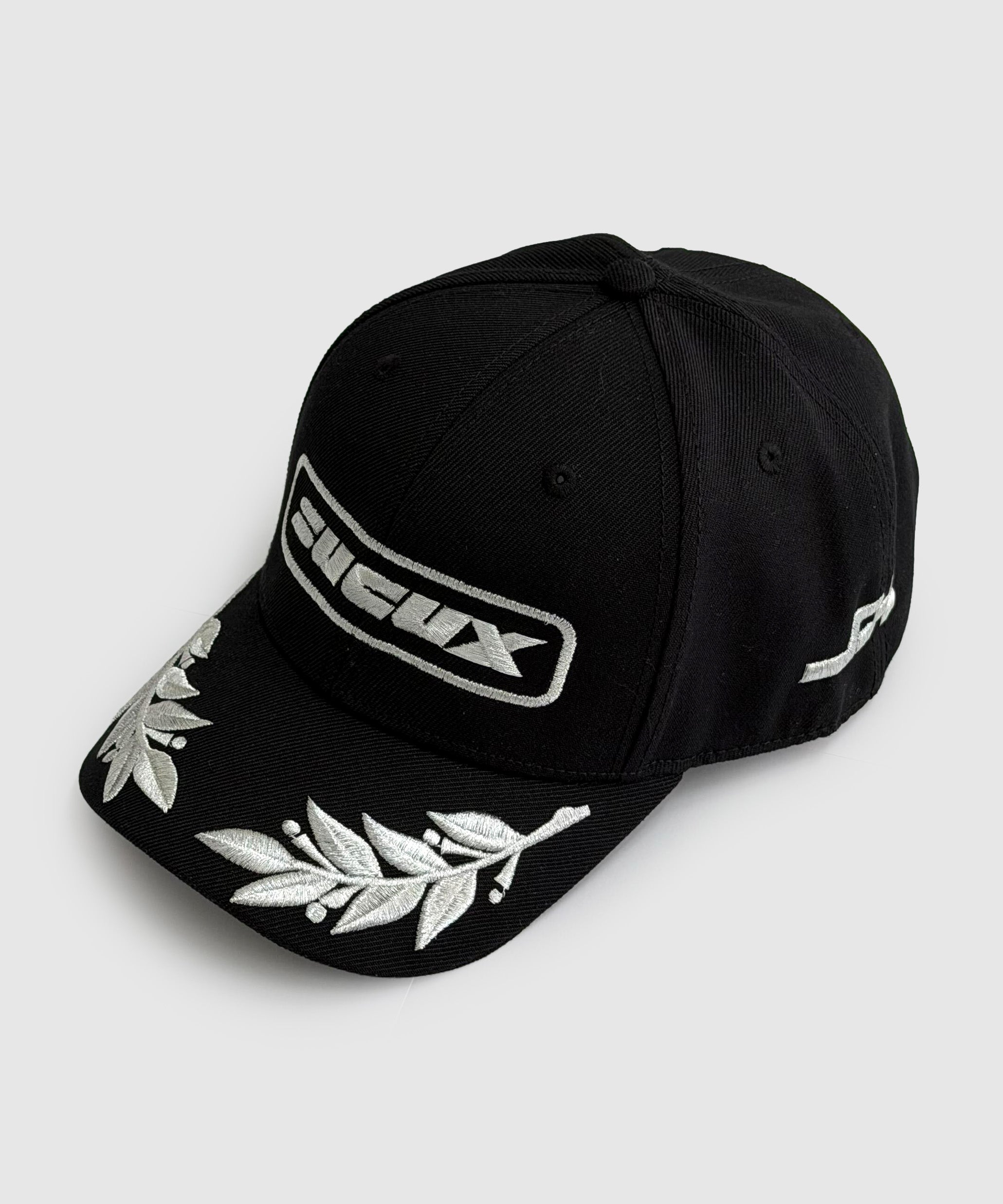 Black baseball cap featuring a silver embroidered “SUCUX” logo on the front, silver laurel leaf embroidery on the brim, and additional white branding on the side.